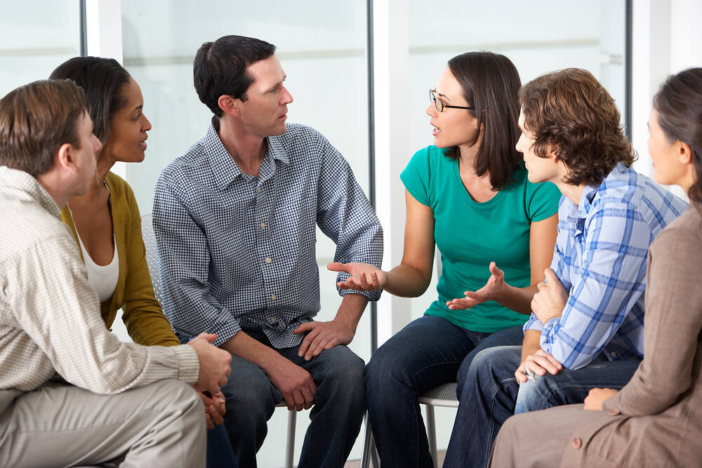 Diverse group of people engaged in a supportive therapy session, discussing addiction recovery in a bright, modern setting