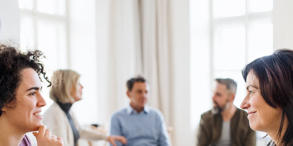 Diverse group of adults engaged in a supportive therapy session for addiction recovery, smiling and interacting in a bright, welcoming room