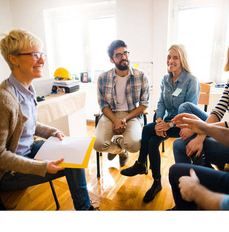 Smiling diverse group of young adults participating in a supportive addiction therapy session, led by a blonde woman with glasses holding a clipboard