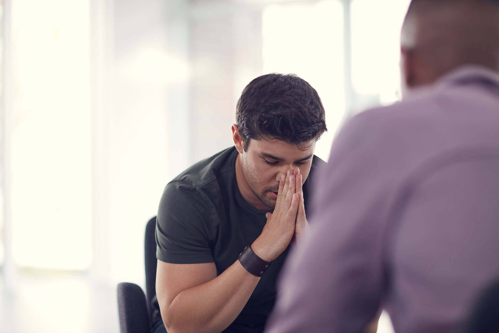 A man sitting in a counseling session, with his head bowed and hands clasped in front of his face, reflecting on his struggles with alcoholism, while another person listens attentively.