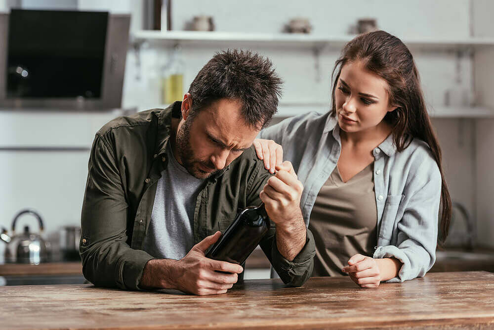 A man holding a bottle of alcohol with a distressed expression, while a woman gently places her hand on his shoulder, illustrating the importance of support in recovery from alcoholism.