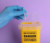 Doctor throwing used syringe needle into sharps container on violet background, closeup